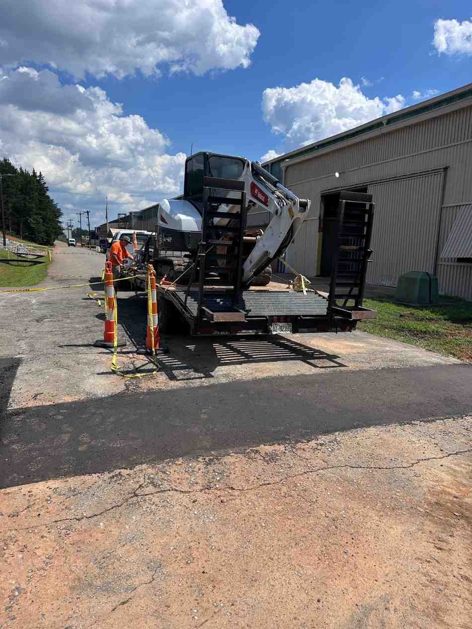 A person in an orange shirt securing a Bobcat skid-steer loader on a trailer next to a building.