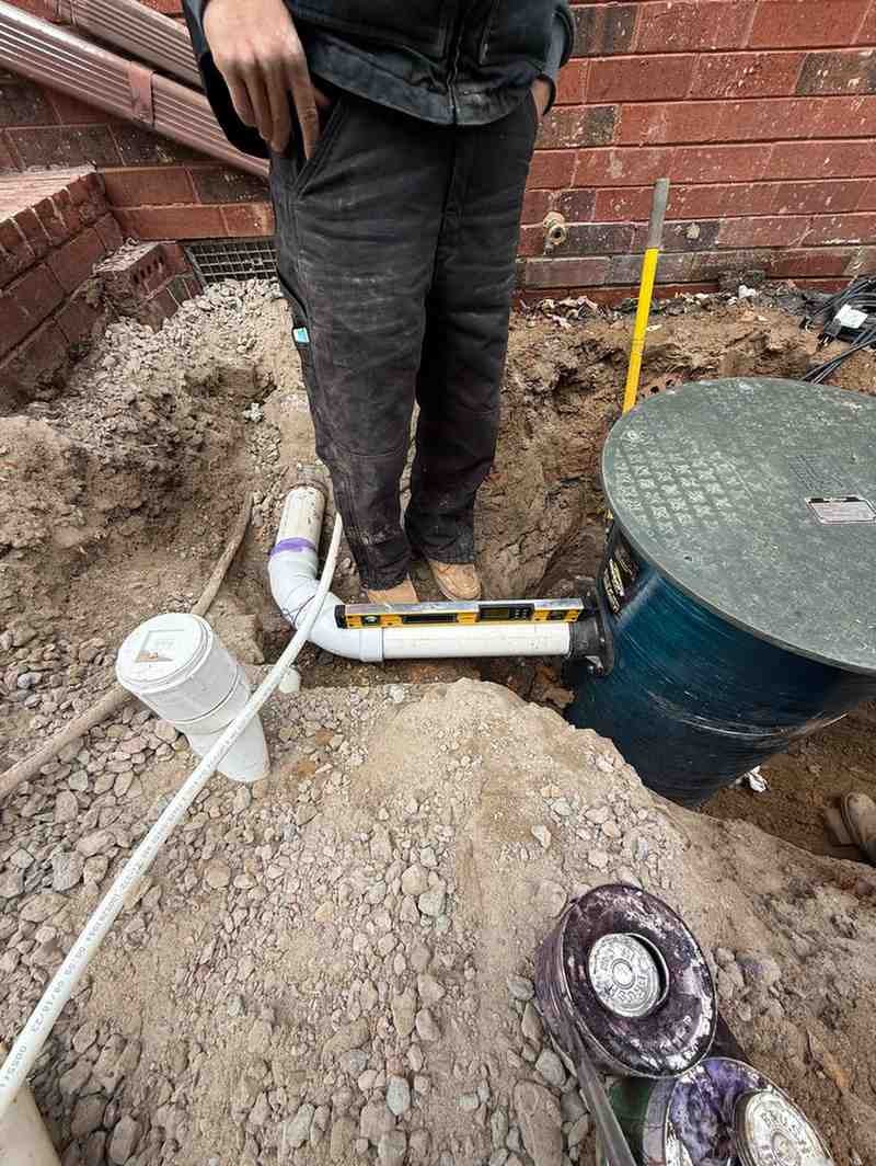 A person works in a trench, laying white PVC pipe with a level, connecting to a black tank, next to bare earth and a brick wall.