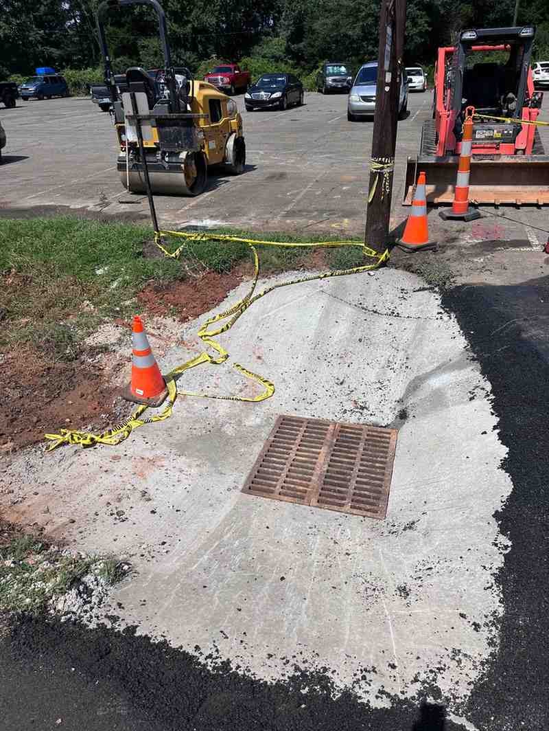 Construction site with a storm drain, fresh concrete, asphalt, caution tape, and vehicles including a paving roller.