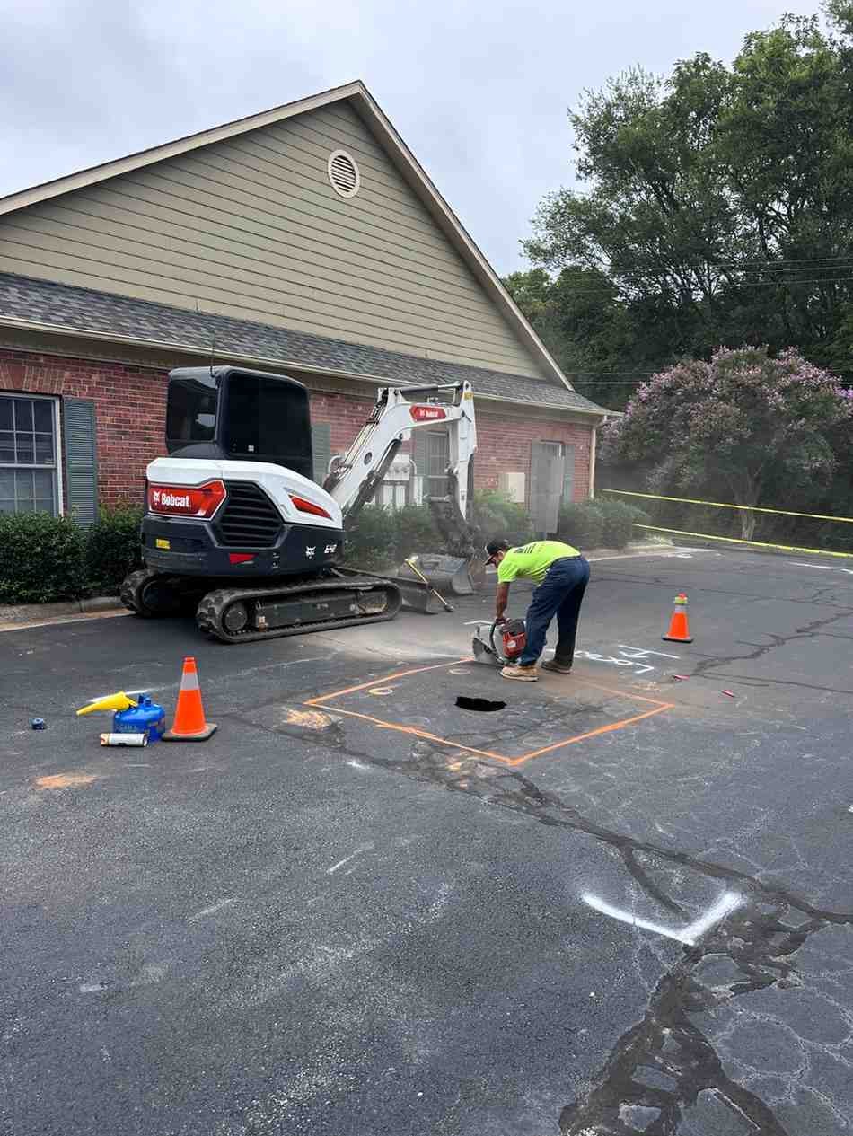 Worker cuts asphalt with a saw near an excavator, orange cone, and building to repair a parking lot.