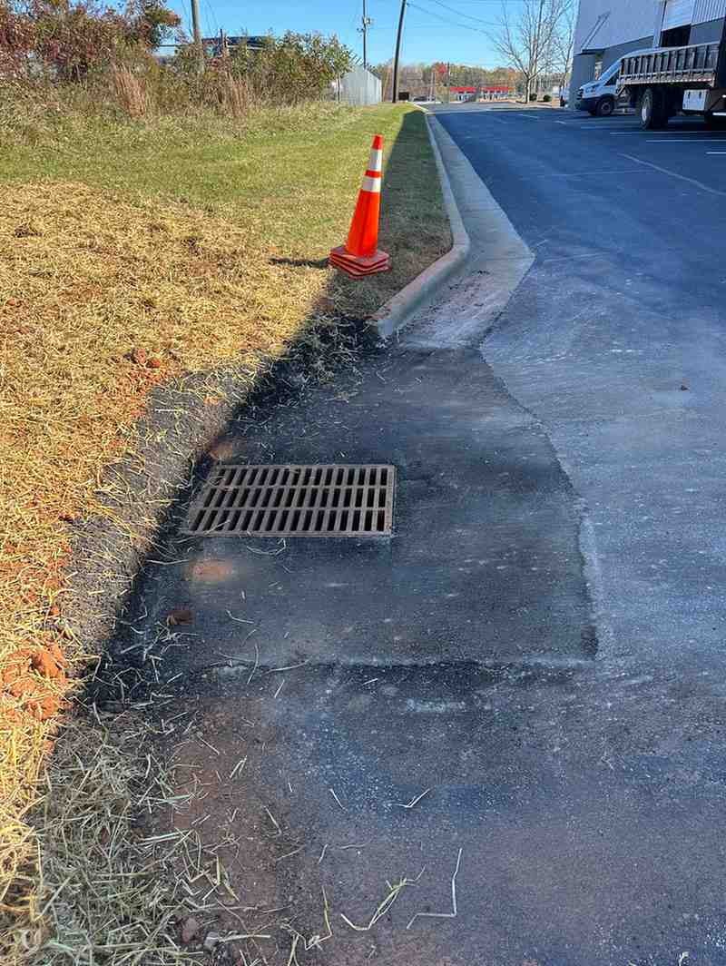 An orange traffic cone stands by a storm drain inlet on an asphalt lot beside a grassy area.
