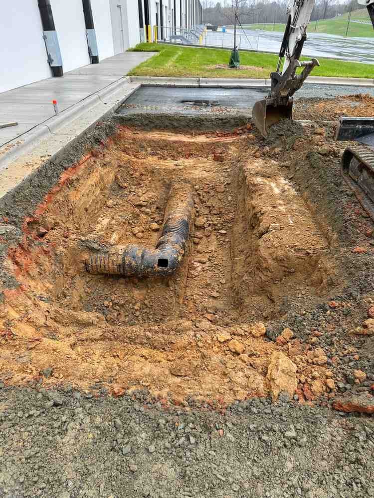 An excavated trench with a black corrugated pipe, next to a building and an excavator bucket.