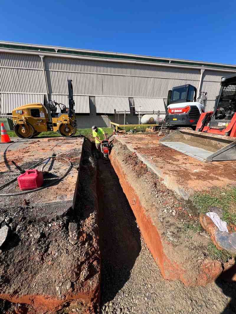 Construction site with a worker digging a trench, surrounded by heavy machinery like a roller and excavator.