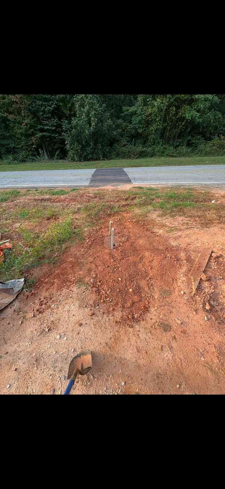 Slightly rusty shovel lying on red dirt, with an asphalt road and dense trees in the background.