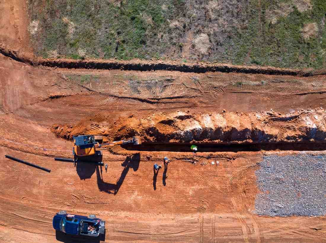 An aerial view of a construction crew digging a long trench in red dirt, with an excavator and a blue truck.