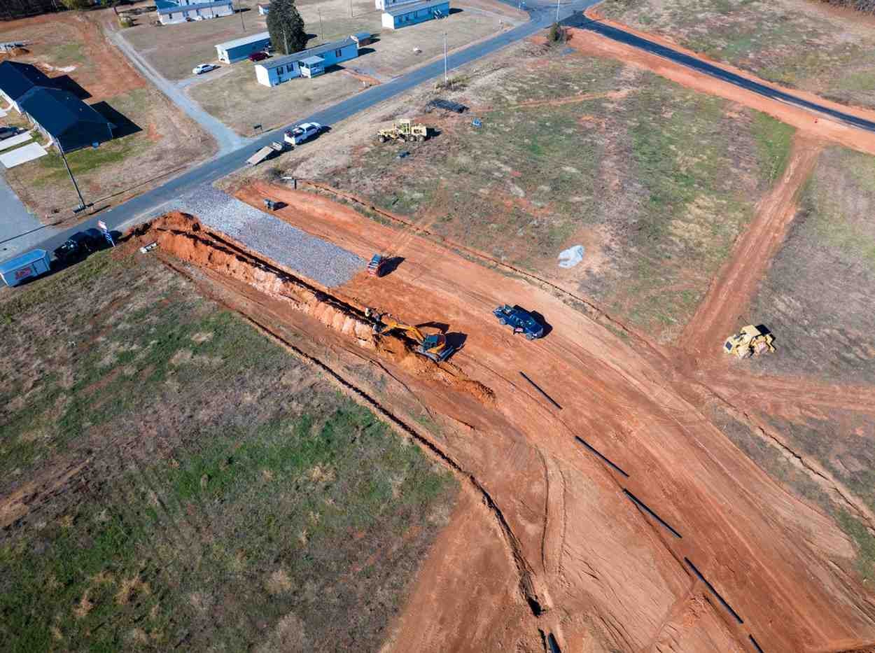 Aerial view of an excavator digging a trench for pipe installation, with construction equipment and dirt roads.