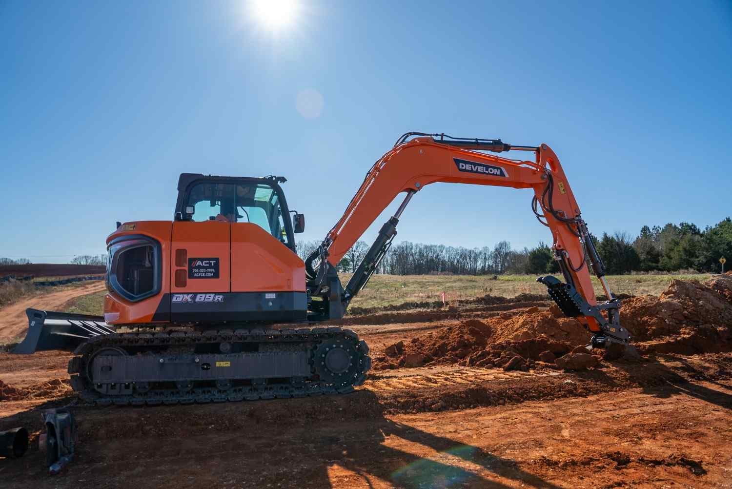 An orange and gray Develon 89R excavator works in a field of red dirt under a bright, sunny blue sky.