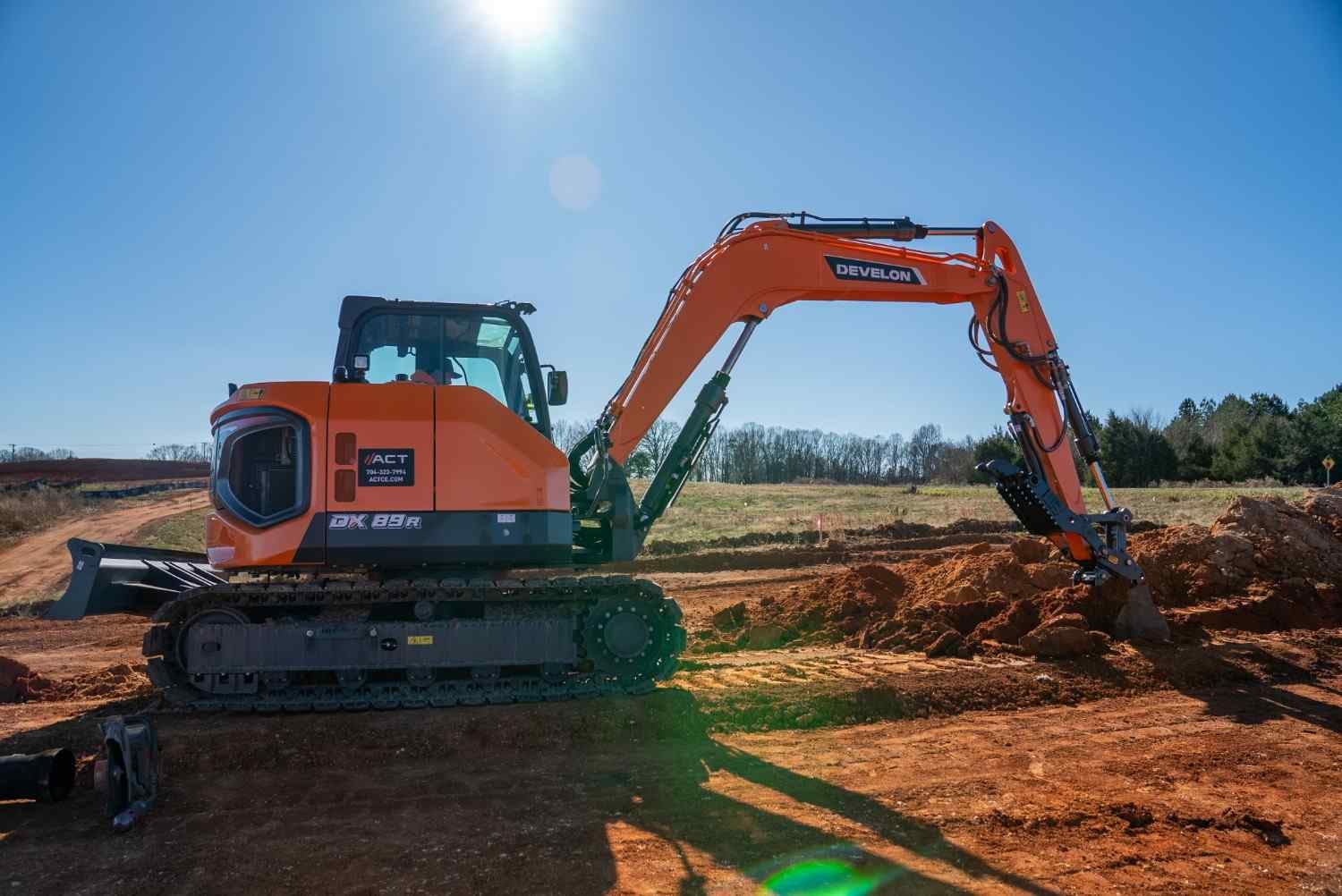An orange Develon DX89R excavator digging into red dirt on a sunny day with trees in the background.