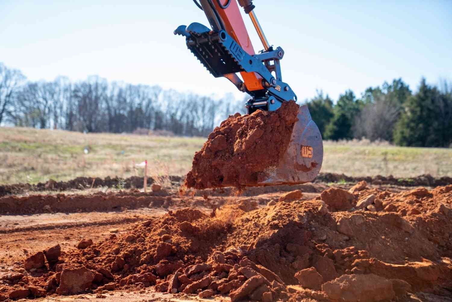 An excavator bucket full of red clay hovers over a pile of dirt, with a field and trees in the background.