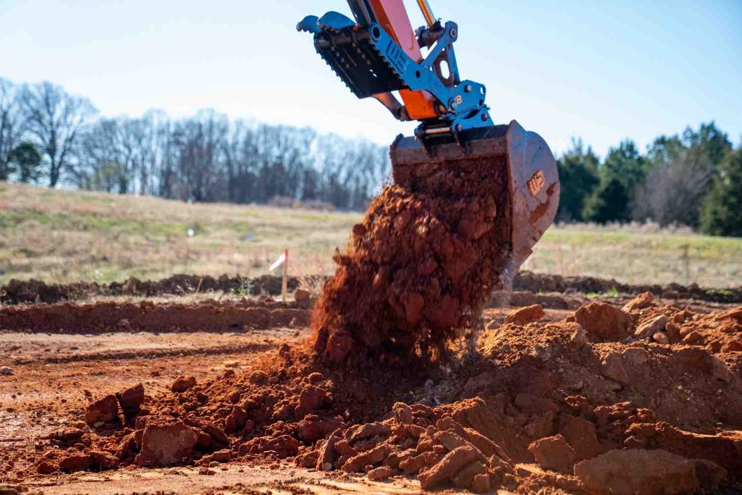 An excavator bucket dumping red soil onto a pile in a field with trees in the background.