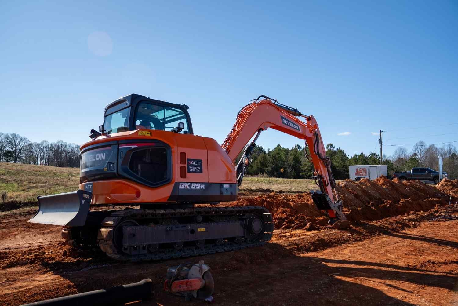 An orange Develon DX89R excavator digging a trench in red dirt under a clear blue sky.