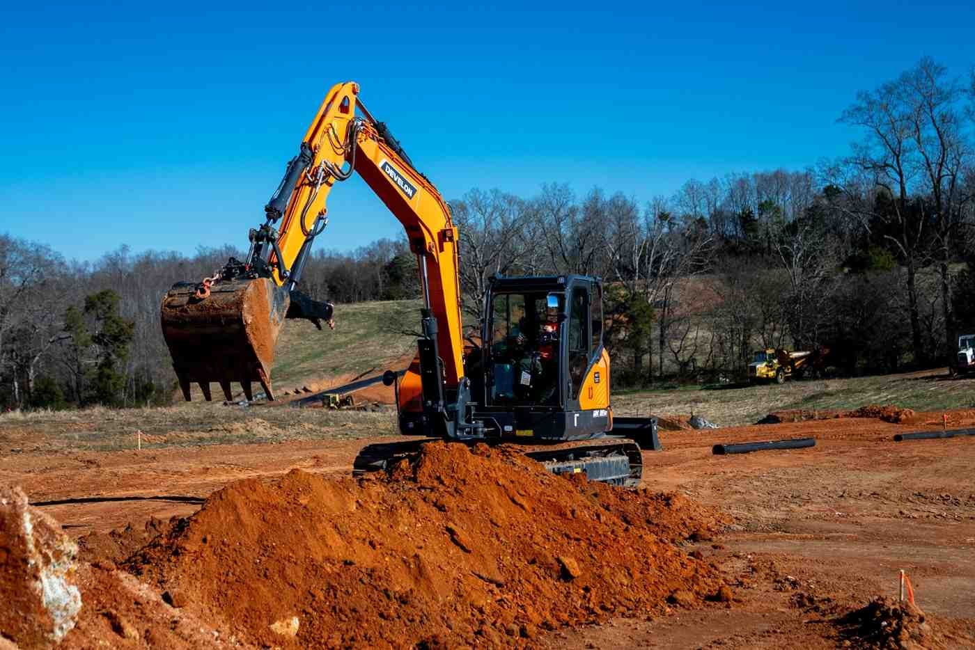 An orange and black excavator with an extended arm and bucket digs an earthen mound into a field.