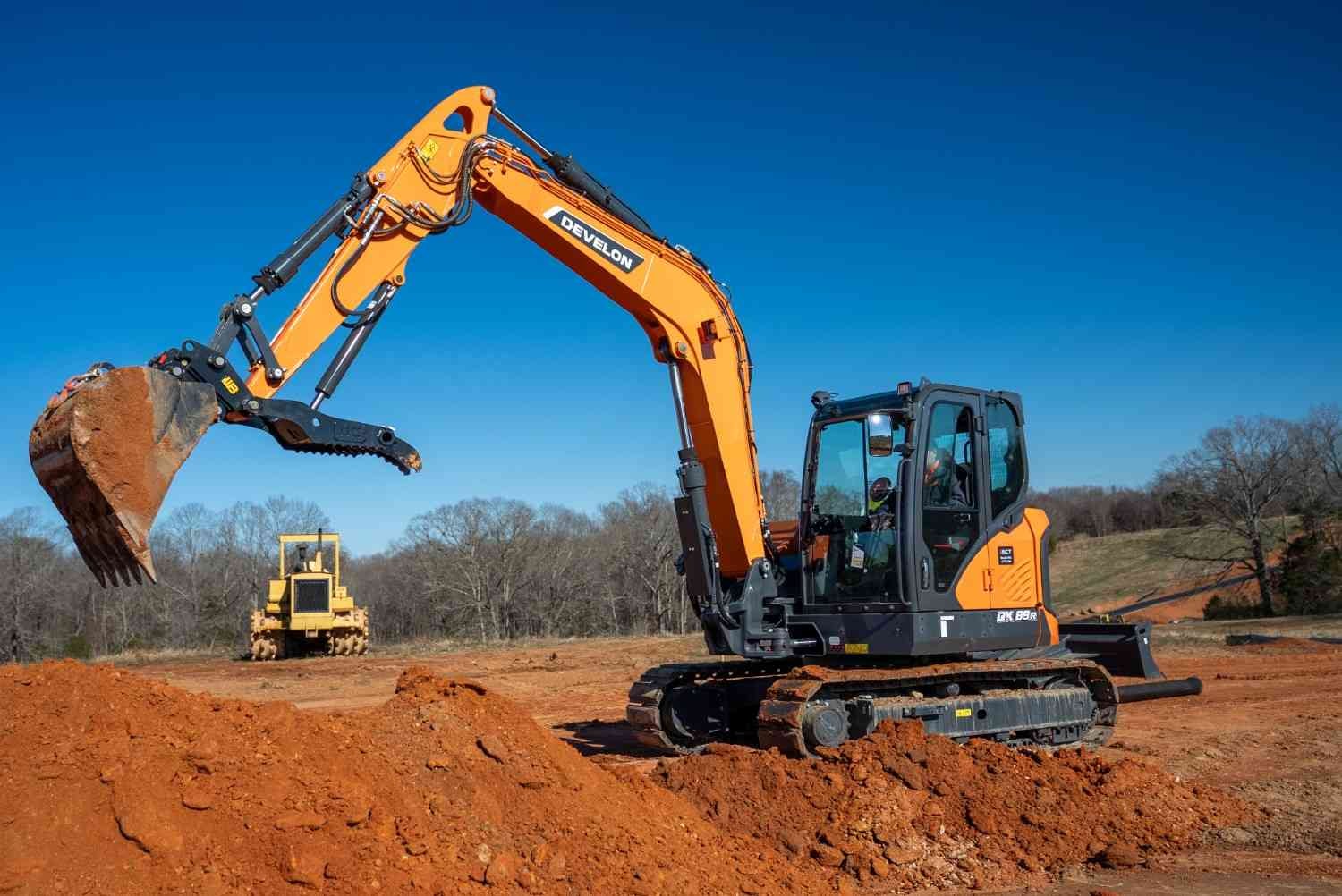 An orange and black Develon excavator pauses with its bucket full of brown dirt on a construction site.