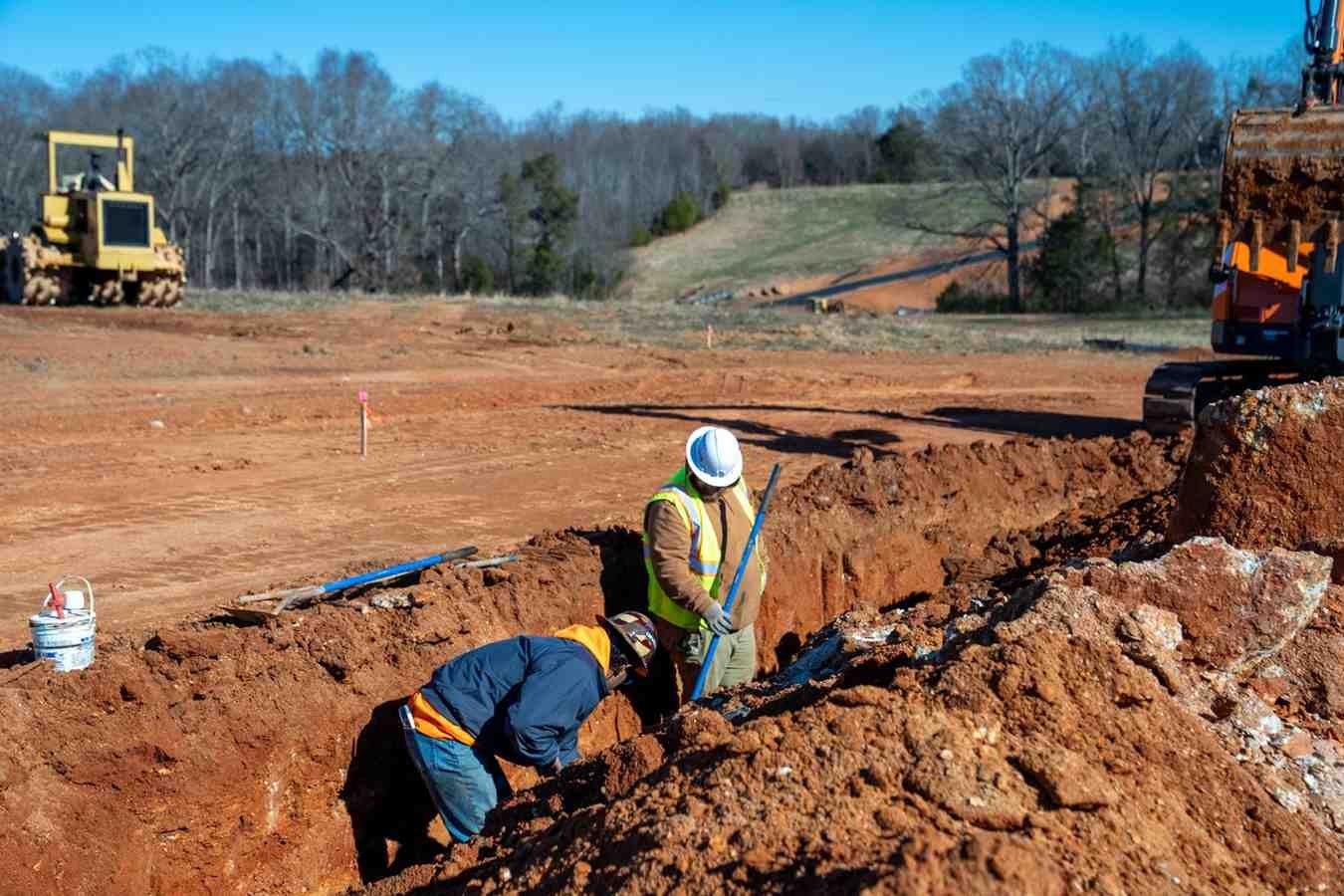 Two construction workers digging a trench on a red dirt site with heavy machinery in the background.