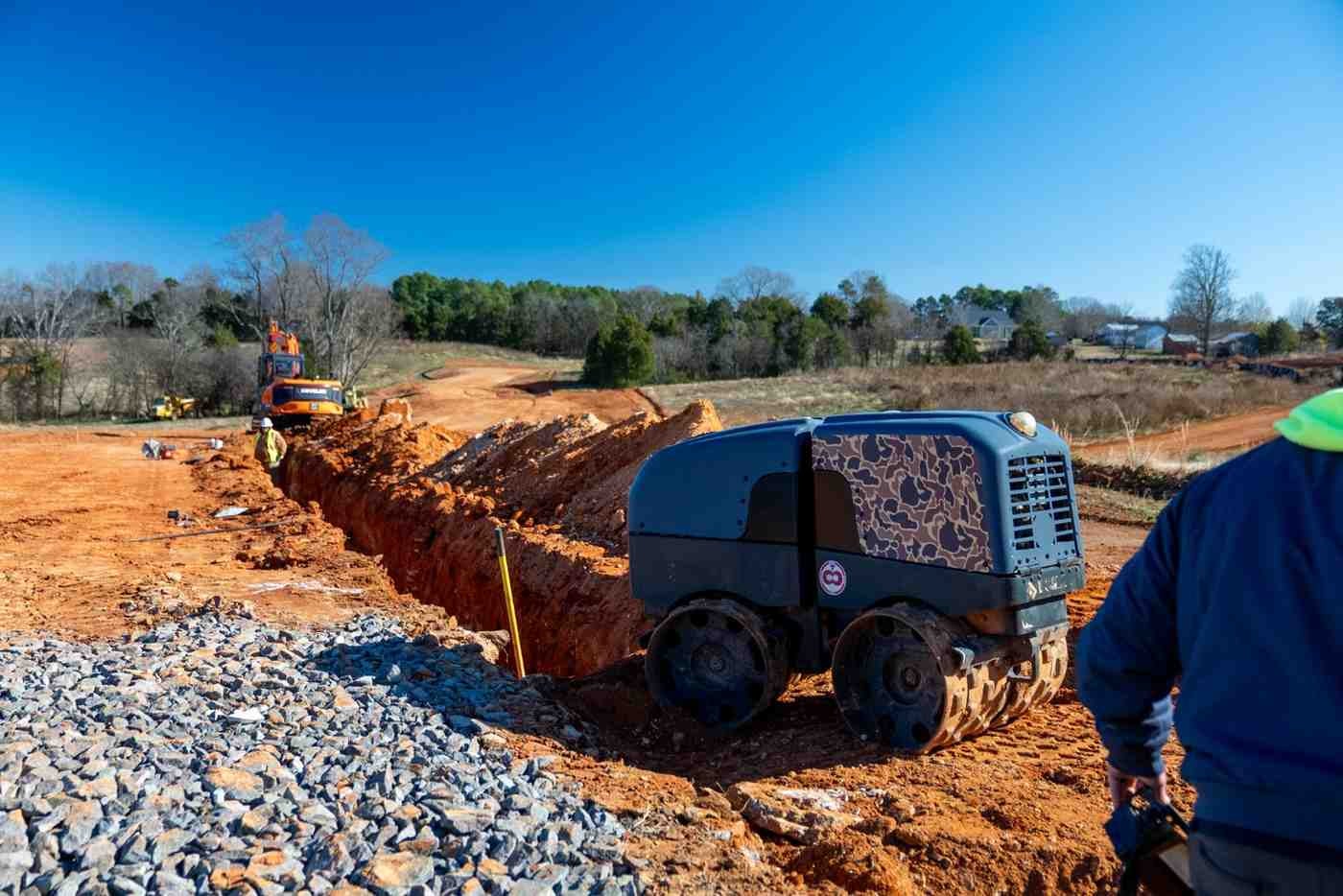 Workers and heavy equipment on a construction site with exposed red dirt, a trench, and gravel under a blue sky.