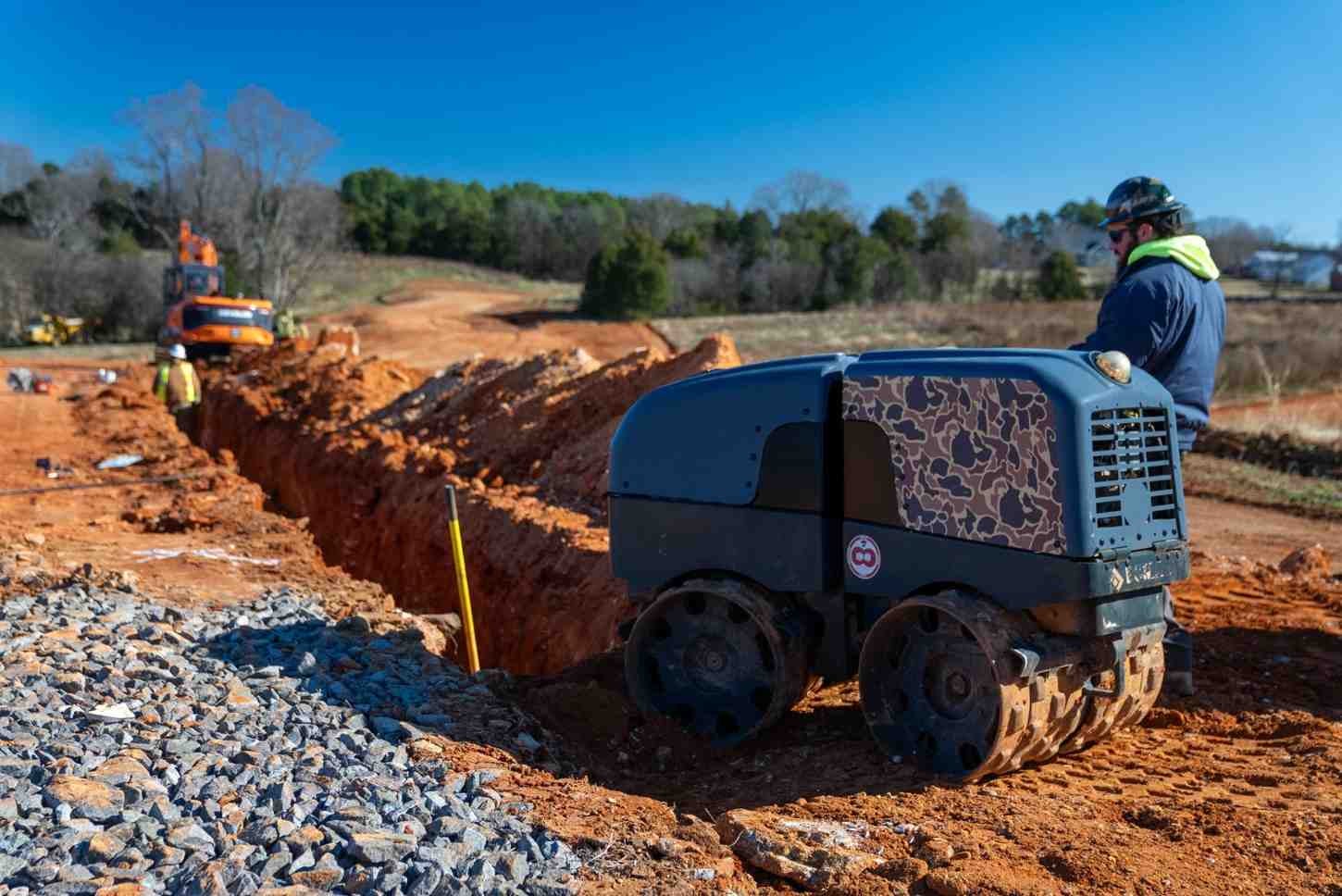 A construction worker in a hardhat operates a vibratory trench roller over red soil near an open trench.