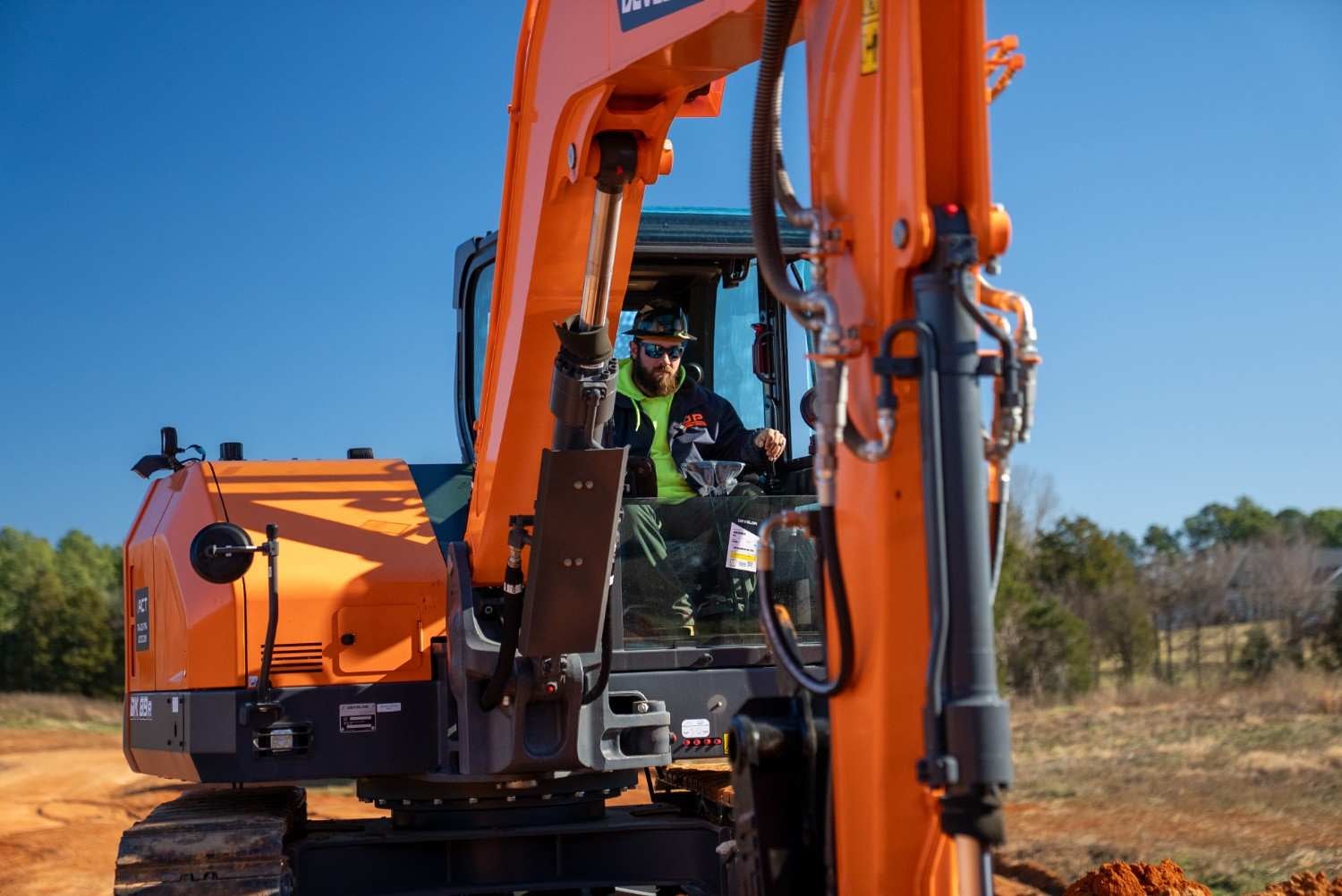 A bearded man in sunglasses, a hard hat, and a jacket operates an orange Develon excavator outdoors.