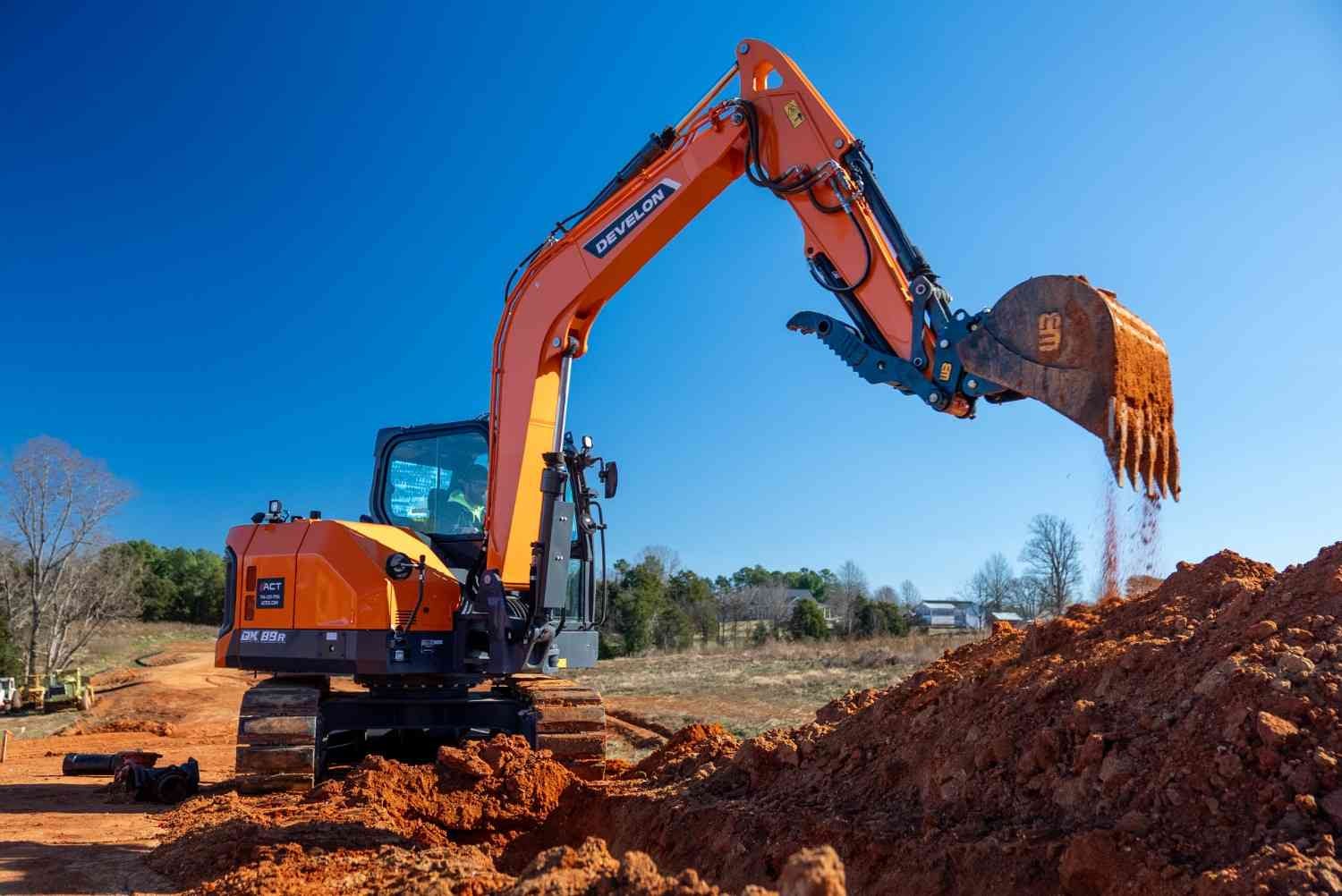 An orange Develon excavator digs into red dirt under a clear blue sky, with trees in the distant background.