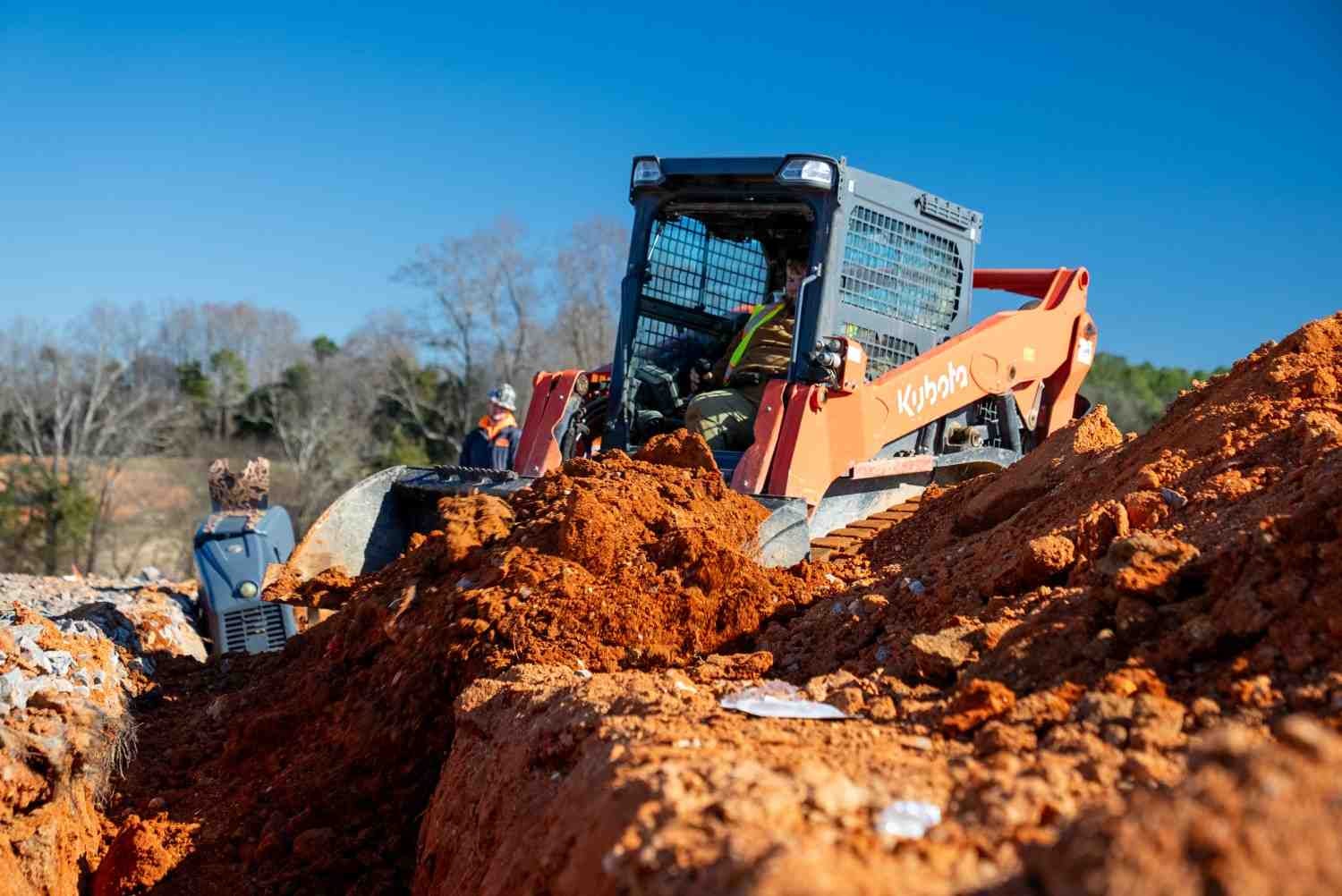 Kubota skid steer moving red dirt on a trench construction site.