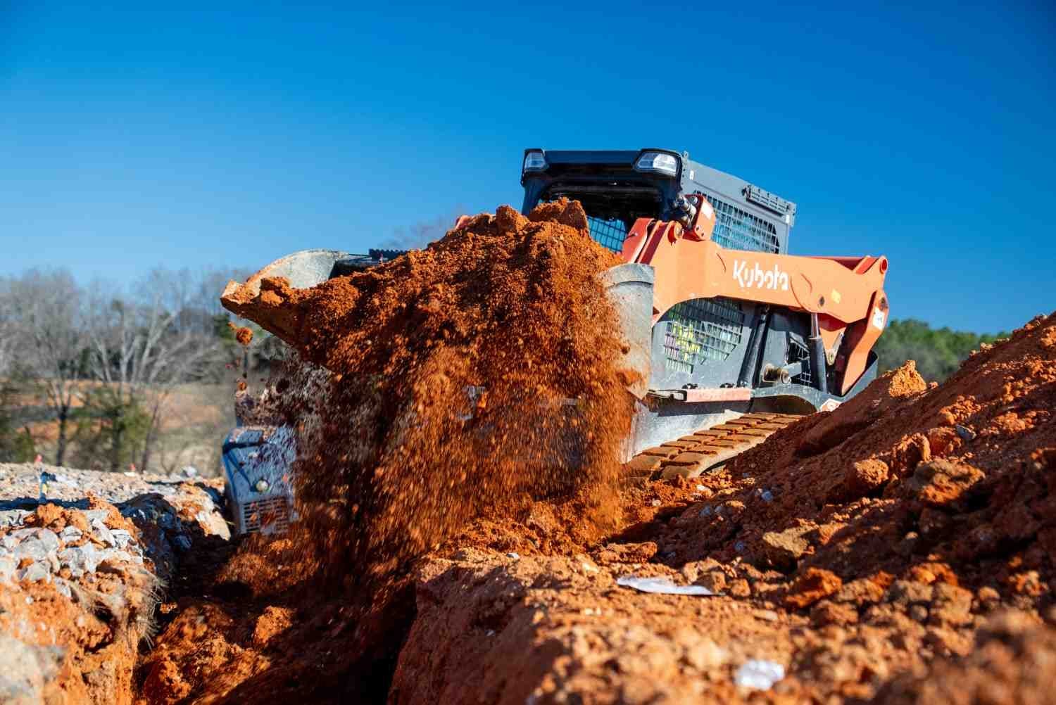 A Kubota skid-steer loader filling a ditch with red earth and rocks on a construction site.