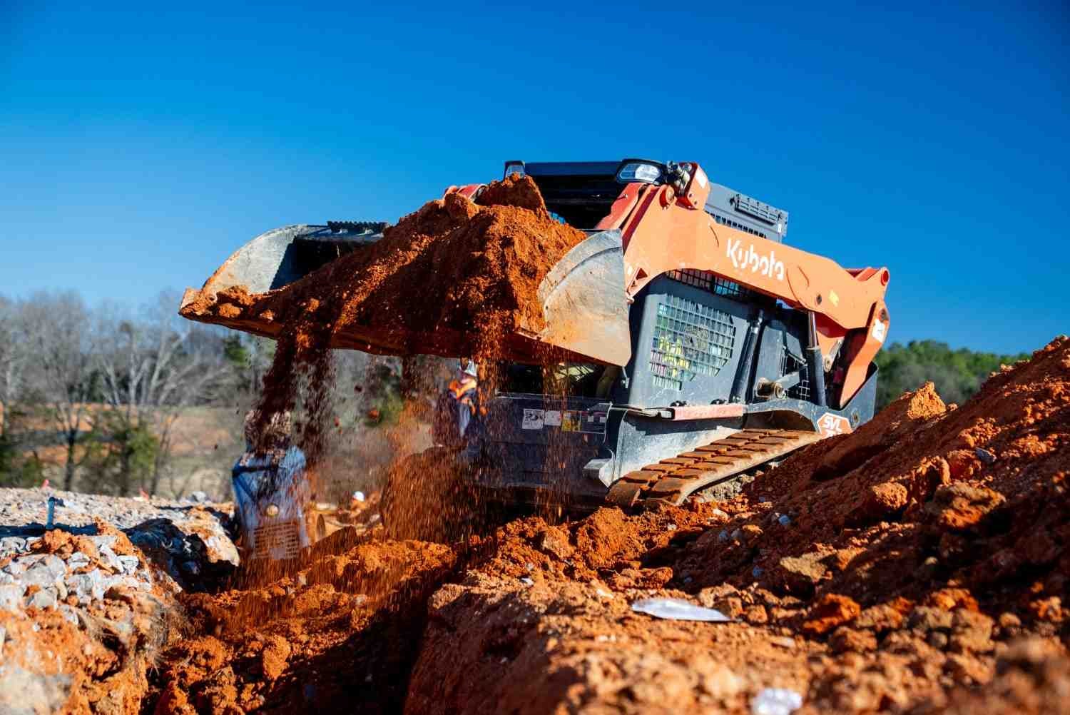 Orange Kubota skid-steer loader dumping a bucketful of red dirt into a trench on a sunny day.
