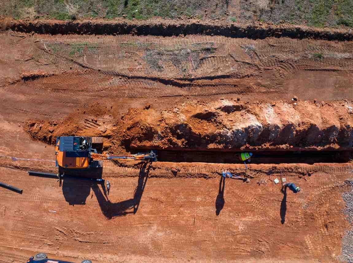Overhead view of an orange excavator digging a trench in reddish earth, with three construction workers.