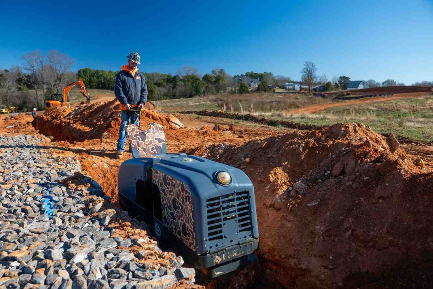 Construction worker operates a remote-controlled trench roller, compacting soil in a dirt ditch with rocks on the left.