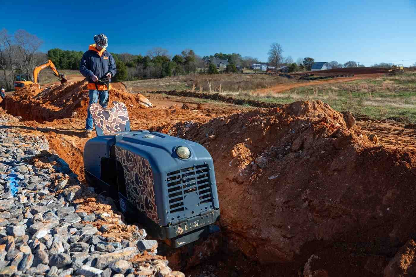 Construction worker operates a remote-controlled trench roller, compacting soil in a dirt ditch with rocks on the left.