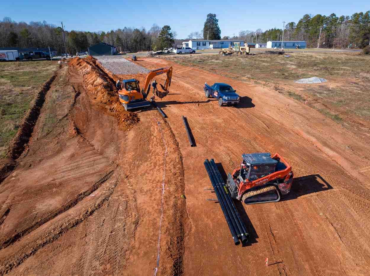 An orange excavator and skid steer with black pipes on a muddy construction site, a blue truck, and buildings in the background.