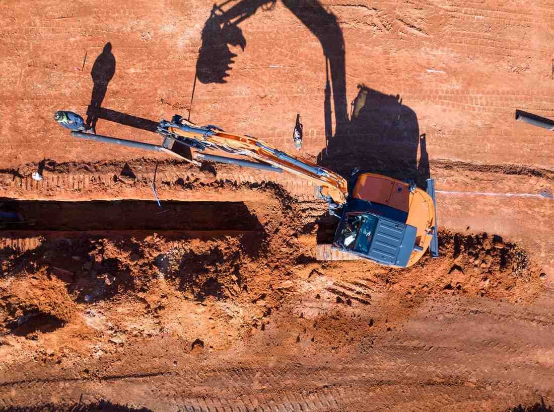 Aerial view of a bright orange excavator parked beside a trench in red dirt, with long shadows.