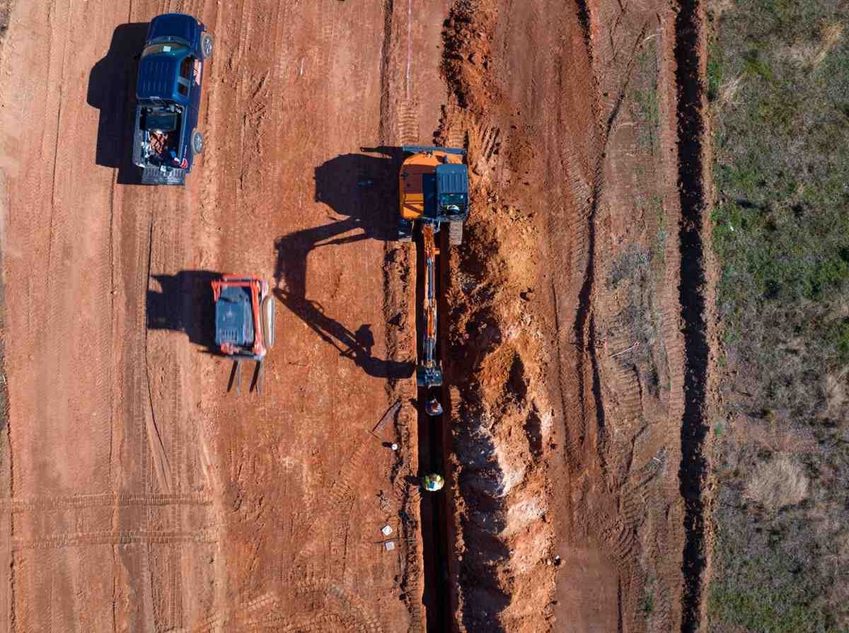 Aerial view of an excavator digging a trench, with two workers, a pickup truck, and a skid steer on red dirt.