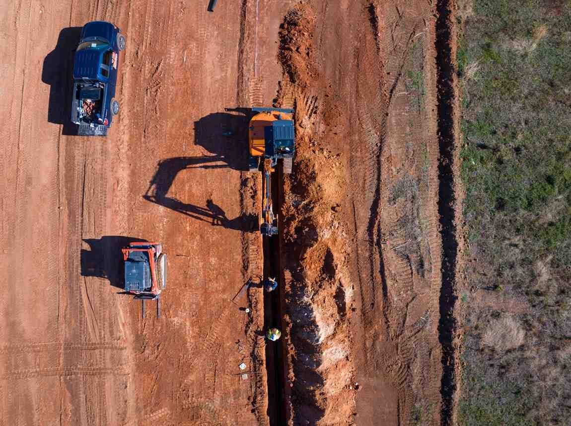 Aerial view of an excavator digging a trench, with two workers, a pickup truck, and a skid steer on red dirt.