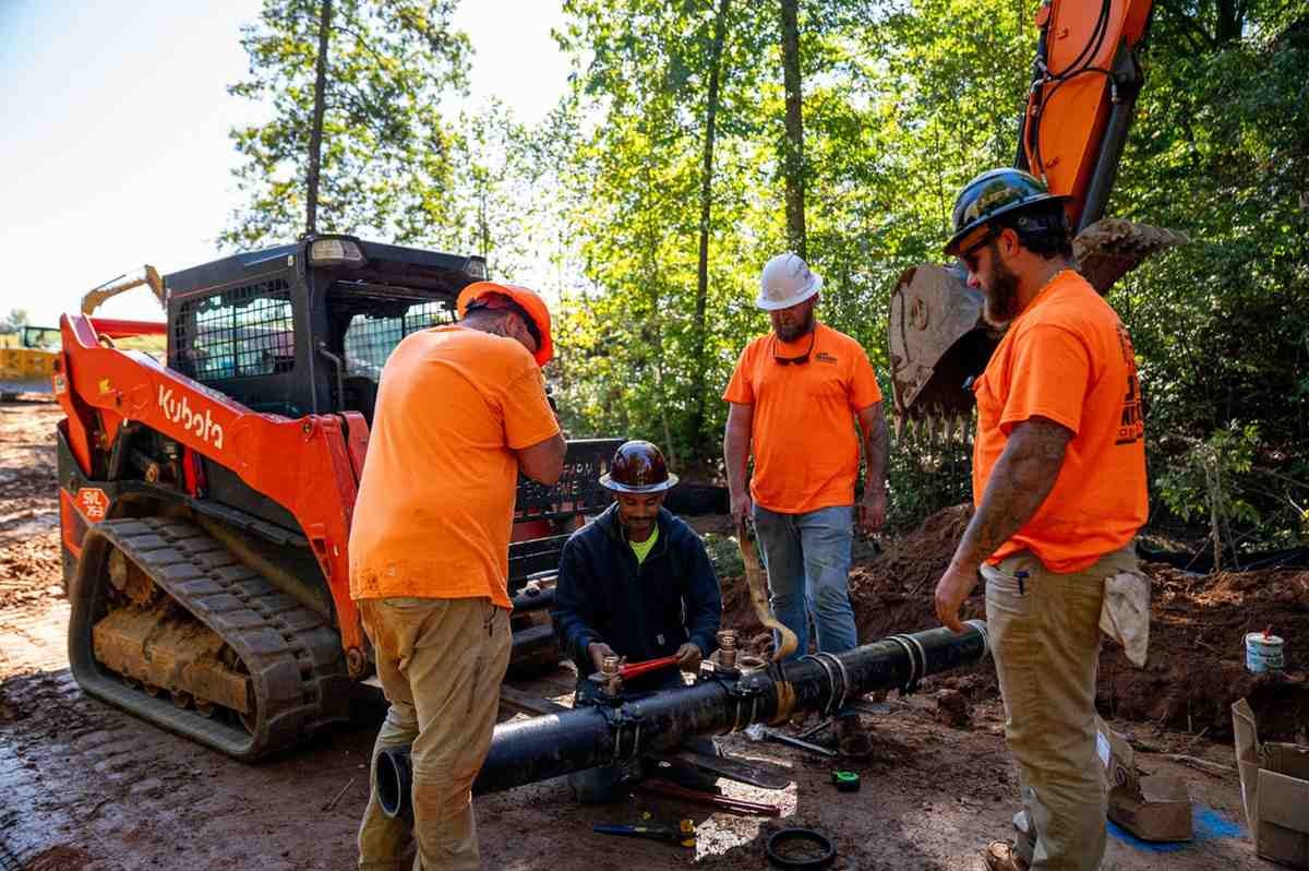 Four construction workers with machinery install a pipe in a wooded area.