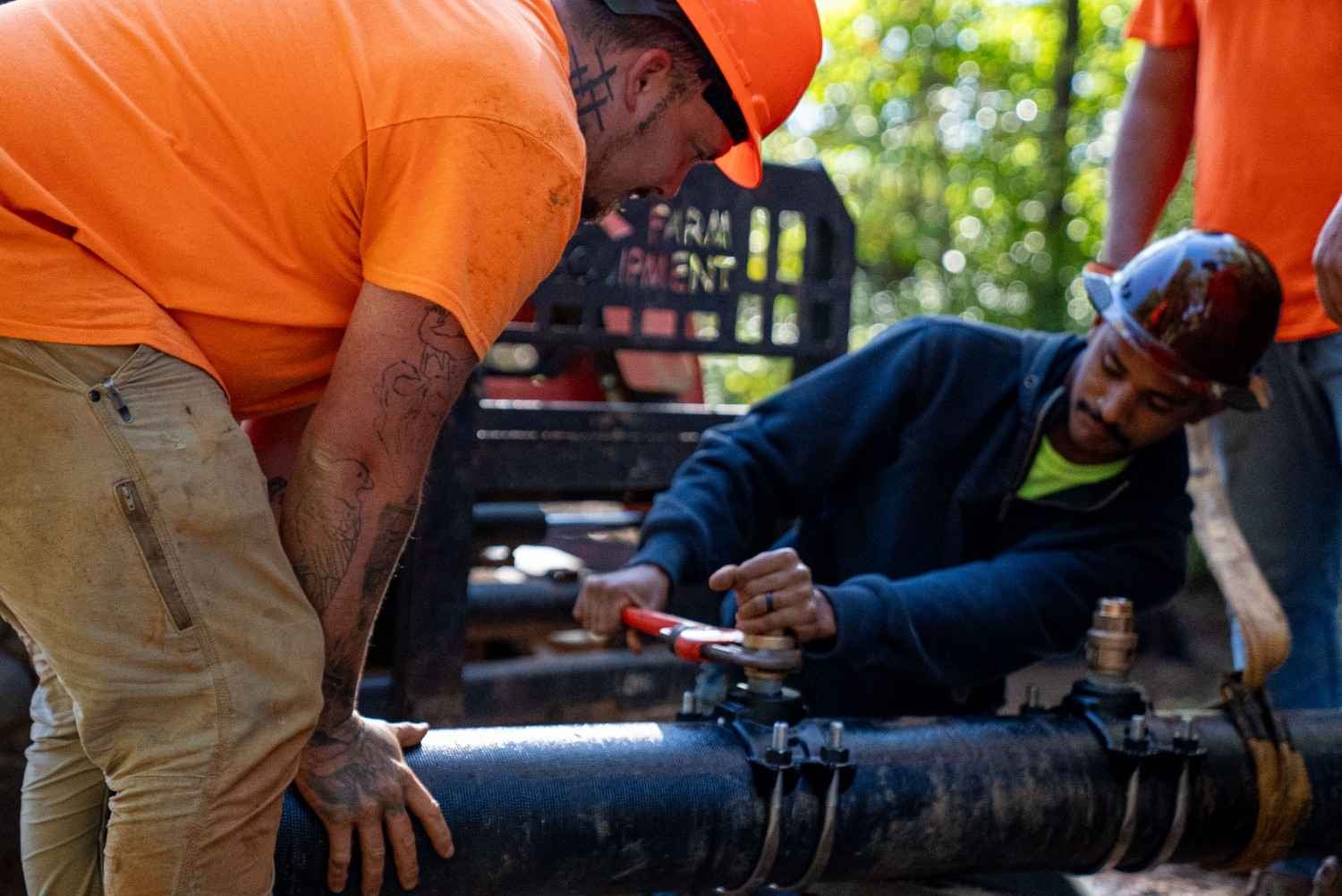 Two construction workers in hard hats connect pipes with a wrench, one bends over, the other watches.