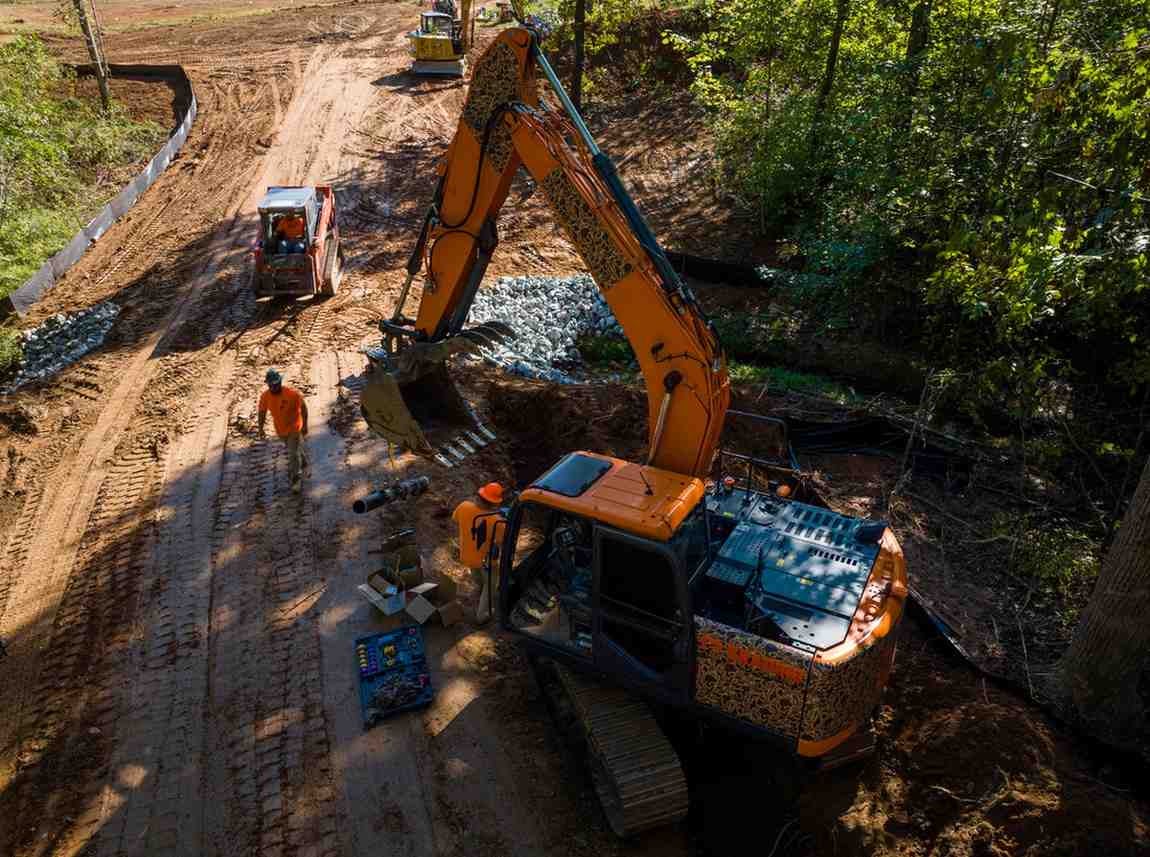Aerial view of construction crew and heavy machinery on a muddy dirt road surrounded by dense woods.