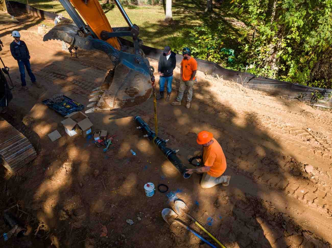 Four construction workers on a dirt road, one kneeling to work on a large pipe next to an excavator.