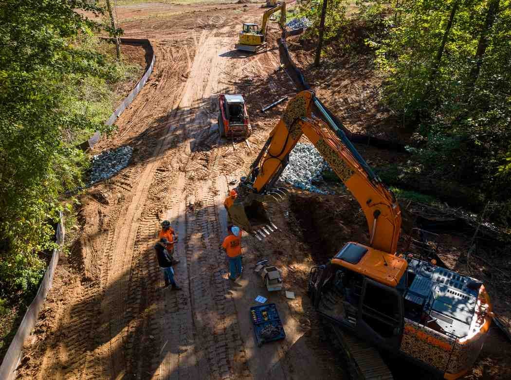 Aerial view of construction crew and heavy machinery on a muddy dirt road surrounded by dense woods.