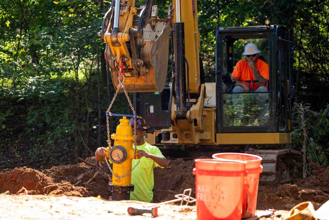 Construction workers install a yellow fire hydrant with an excavator in a wooded, muddy area.