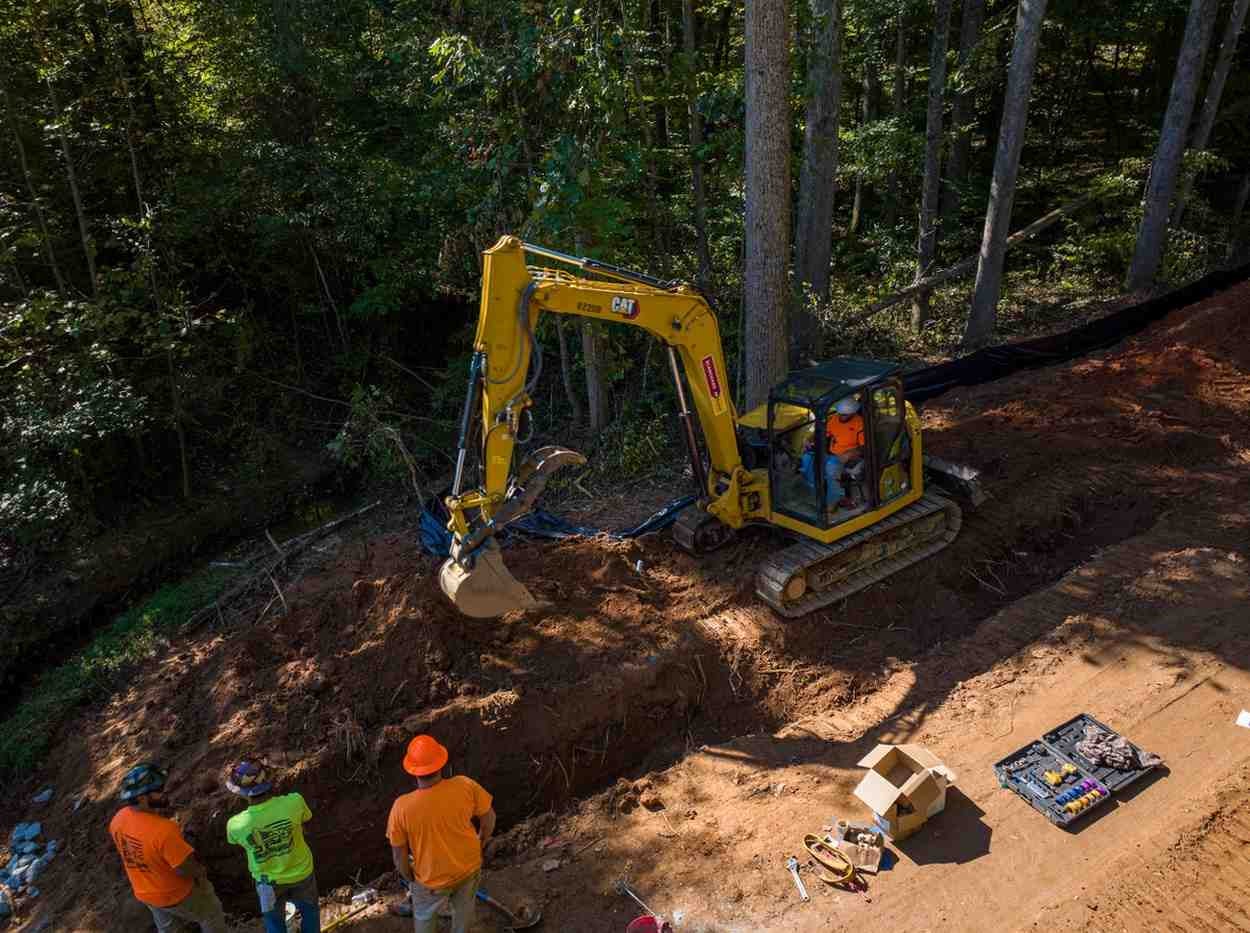 Aerial view of a yellow excavator digging a trench, with three workers observing from below near woods.