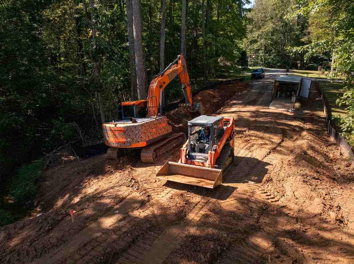 An orange excavator and skid steer on a dirt construction site, surrounded by green trees under a sunny sky.