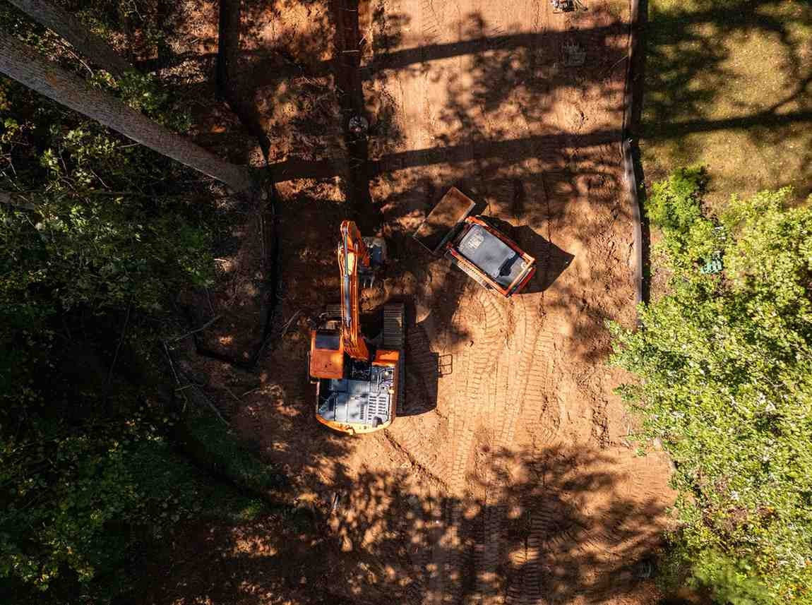 Aerial view of an orange excavator and small skid-steer loader on a brown construction site between green trees.