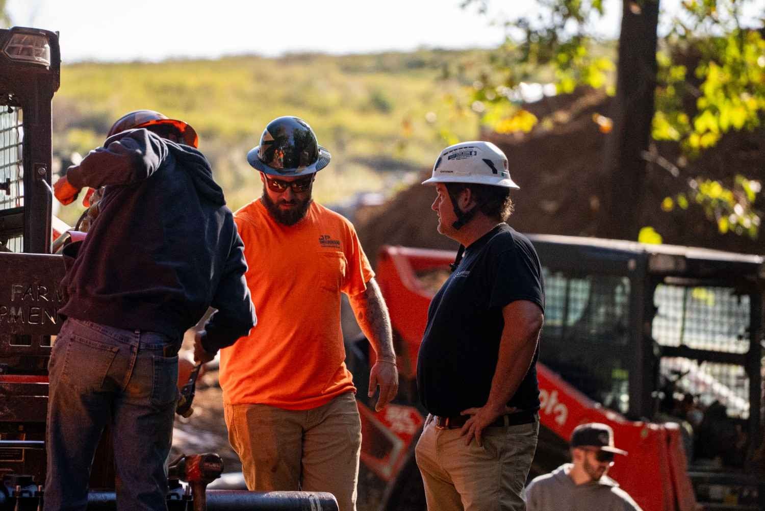 Three construction workers in hard hats and work clothes, two looking at each other, on a job site with machinery.