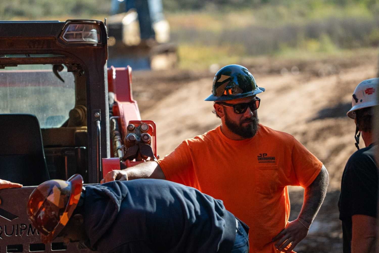 A bearded man in an orange shirt, hard hat, and sunglasses observes another worker near heavy machinery.