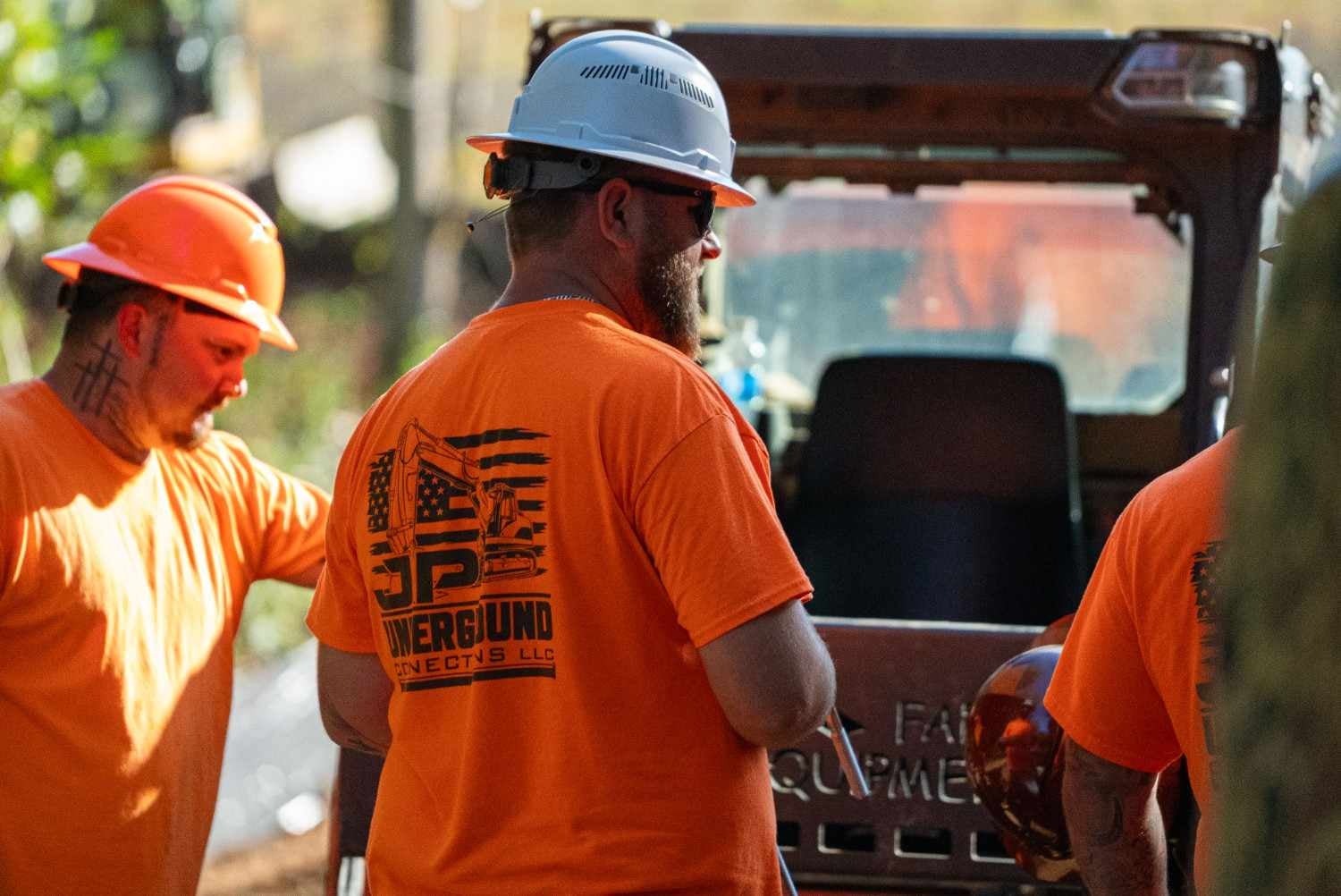 Three construction workers in orange shirts and hard hats, two with their backs to the camera, next to a vehicle.