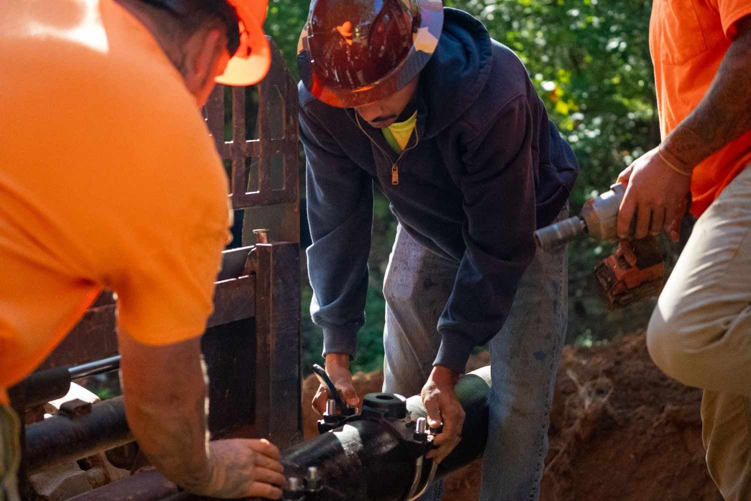 Three construction workers in hard hats and bright shirts installing a black pipe.